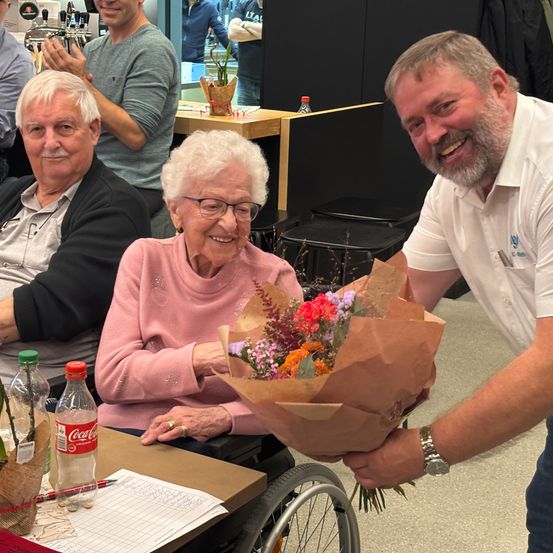Ein Mann überreicht einer älteren Frau im Rollstuhl einen Blumenstrauß, während andere hinter ihnen in einem Raum klatschen.