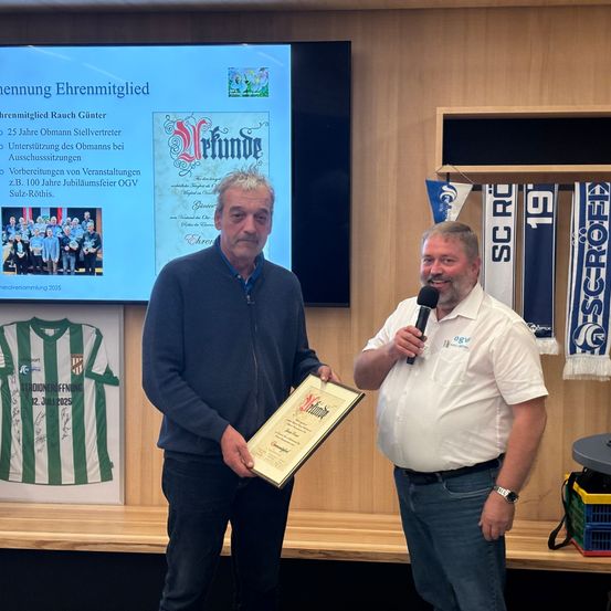 Zwei Männer stehen vor einer Holzwand mit einem gerahmten Zertifikat, einer hält es und der andere ein Mikrofon. Dahinter befindet sich eine Projektionsleinwand mit einem Gruppenbild und Text. Ein Trikot und Banner sind ebenfalls zu sehen.