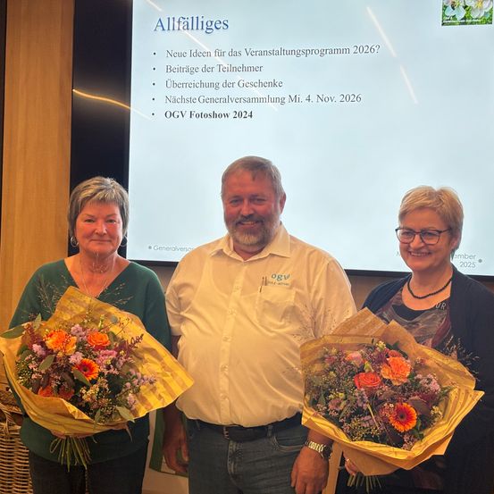 Drei Menschen stehen lächelnd vor einer Leinwand, zwei Frauen halten Blumensträuße, eine in Grün, die andere in Schwarz. Der Mann in Weiß trägt eine Uhr. Dahinter zeigt eine Leinwand den Text 'Allfalliges'.