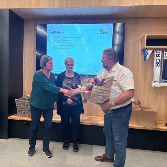 Drei Personen stehen in einem Raum, zwei Frauen und ein Mann. Der Mann hält einen Blumenstrauß und schüttelt die Hände der Frauen. Eine Projektionsleinwand im Hintergrund zeigt eine Präsentation.