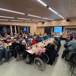 Eine große Gruppe von Menschen versammelt sich in einem Raum, sitzt an Tischen, mit einer Person, die vorne steht. Der Raum hat Holzwände und eine Projektionsleinwand. Die Menschen interagieren miteinander.