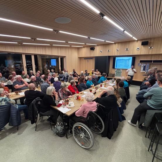Eine große Gruppe von Menschen versammelt sich in einem Raum, sitzt an Tischen, mit einer Person, die vorne steht. Der Raum hat Holzwände und eine Projektionsleinwand. Die Menschen interagieren miteinander.