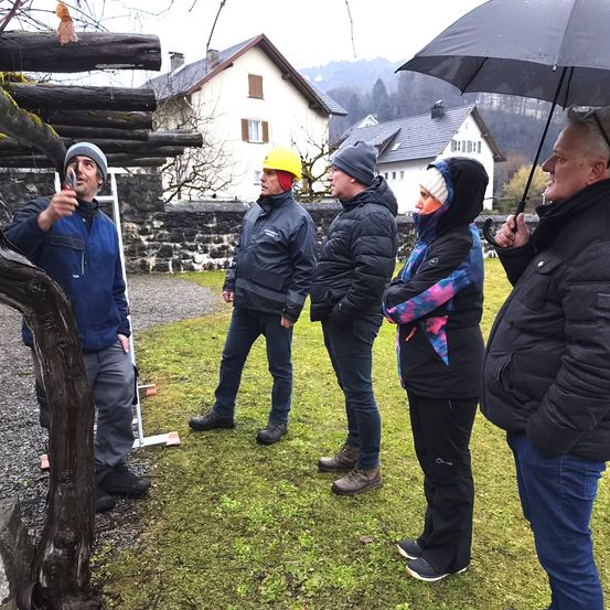 Fünf Personen stehen draußen, eine von ihnen hält ein Werkzeug. Sie tragen Jacken und einige Mützen. Sie stehen vor einem Haus mit Schnee auf dem Dach. Der Boden ist grasig.