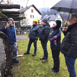 Fünf Personen stehen draußen, eine von ihnen hält ein Werkzeug. Sie tragen Jacken und einige Mützen. Sie stehen vor einem Haus mit Schnee auf dem Dach. Der Boden ist grasig.