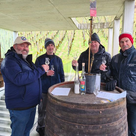 Vier Männer stehen um einen Fass in einer Außenumgebung herum, alle halten Weingläser. Hinter ihnen ist ein Weinberg sichtbar, und sie scheinen Wein zu diskutieren oder zu verkosten.