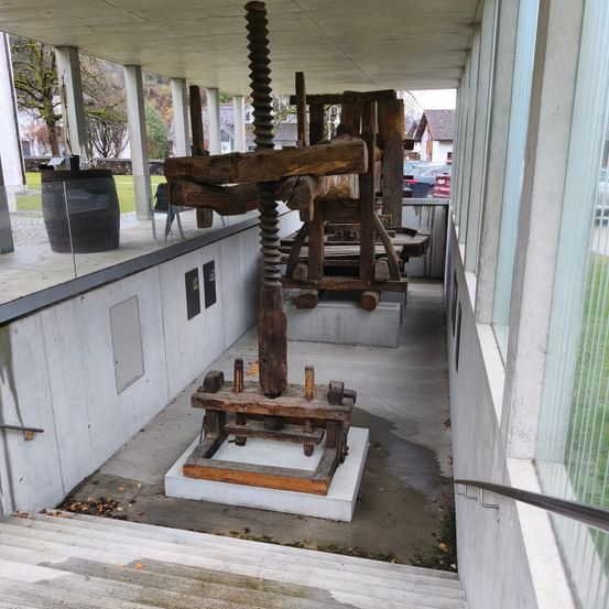 Eine alte, große Holzpresse ist auf einer Betonplattform in einem modernen Gebäude ausgestellt. Die Presse ist von Betonbarrieren umgeben. In der Nähe steht ein großer Fass und ein Gebäude mit einem roten Auto, das davor geparkt ist.