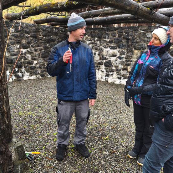 Ein Mann in einem blauen Jackett erklärt etwas zwei Frauen in Winterkleidung, die draußen in einem Schotterbereich stehen.