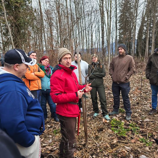 Eine Gruppe von Menschen in Winterkleidung steht in einem Wald und hört einem Mann in einer roten Jacke zu, der einen Stock hält.