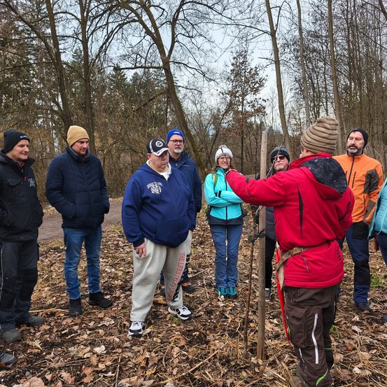 Eine Gruppe von Erwachsenen steht in einem Wald. Ein Mann in einem roten Mantel hält einen Stock und scheint der Gruppe etwas zu erklären. Die Gruppe hört aufmerksam zu.