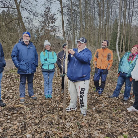 Eine Gruppe von Menschen steht in einem Wald. Ein Mann in einem blauen Mantel hält einen Stock. Sie tragen alle Winterkleidung. Sie halten wahrscheinlich eine Versammlung ab.