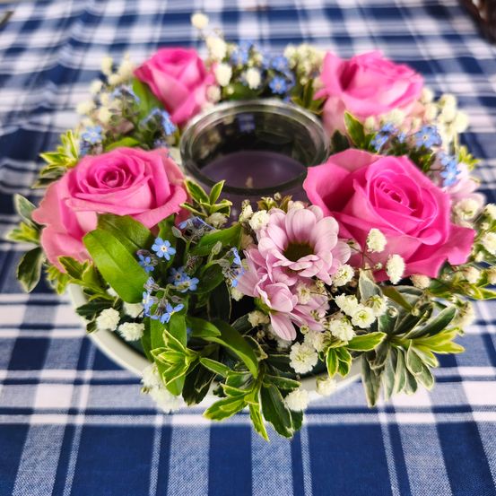 Ein Gesteck mit rosa Rosen, blauen Blumen und grünen Blättern auf einem blau-weiß karierten Tischtuch, mit einer Kerze in der Mitte.