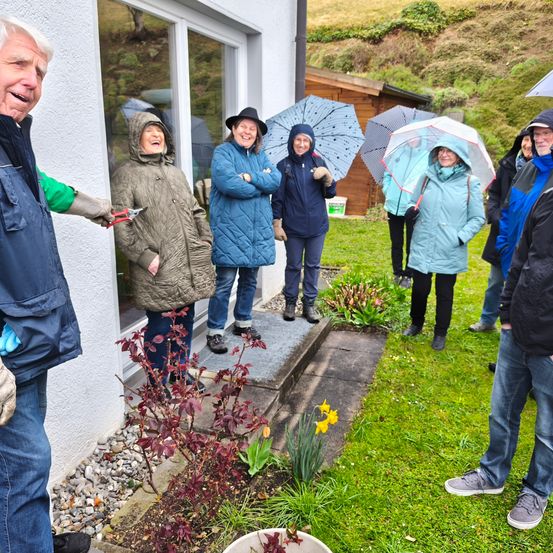 Eine Gruppe von Menschen in Regenmänteln und Handschuhen steht in einem Garten mit Regenschirmen. Eine Person lächelt und hält eine Schere in der Hand.