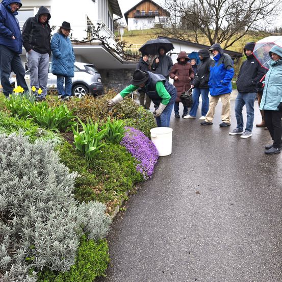 Eine Gruppe von Menschen in Regenkleidung ist um einen Garten versammelt, wobei eine Person sich um Blumen kümmert. Die Szene spielt sich in einer regnerischen Umgebung ab, mit einem Gebäude und einem geparkten Auto im Hintergrund.