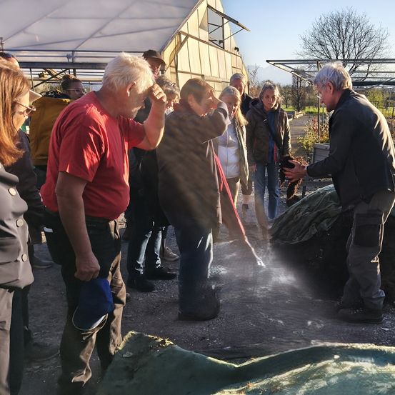 Eine Gruppe von Menschen, einschließlich eines Mannes in Rot, steht in einem Außenbereich, möglicherweise in einem Garten. Ein Mann in Schwarz hält eine Katze, und die Menschen scheinen die Katze anzuschauen.
