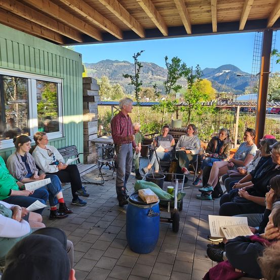 Ein Mann steht vor einer Gruppe von Menschen, die auf einer Bank sitzen und ein Buch lesen. Hinter ihnen befinden sich Pflanzen, Bäume und Berge.