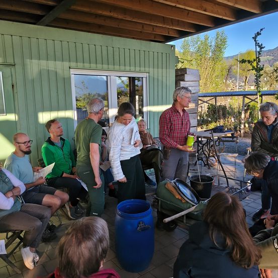 Eine Gruppe von Menschen in einem Meeting, stehend und sitzend um einen Raum herum. Ein Mann hält einen Becher, während eine Frau eine Mappe in der Hand hat. Es gibt Fässer und Tische im Raum.