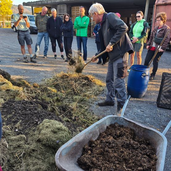 Ein älterer Mann benutzt eine Schaufel, um Kompost in eine Schubkarre zu schaufeln, während mehrere andere zusehen.