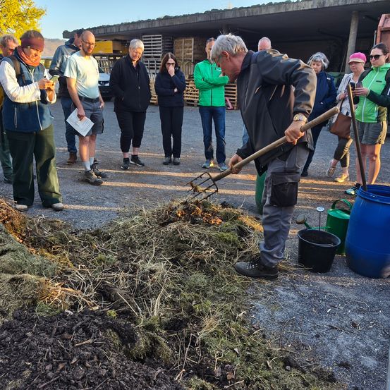 Eine Gruppe von Menschen steht um einen Mann mit einer Heugabel herum. Er scheint den Boden zu hacken. Es gibt Eimer und Fässer um ihn herum. Die Leute beobachten ihn.