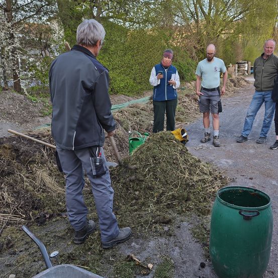 Eine Gruppe von Menschen ist im Freien. Ein Mann gräbt mit einer Schaufel, während ein anderer Mann auf einem Telefon schreibt. Eine Frau steht mit einem grünen Eimer in der Nähe.