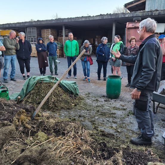 Eine Gruppe von Menschen ist um einen Mann versammelt, der etwas erklärt. Sie sind draußen in der Nähe eines Haufens Erde und eines großen grünen Fasses.