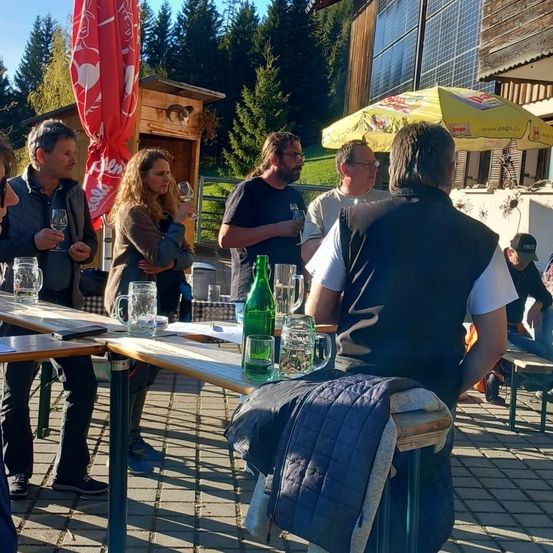 Eine Gruppe Erwachsener versammelt sich vor einem Restaurant, einige mit Weingläsern, unter einem gelben Regenschirm. Bäume und ein kleines Gebäude sind im Hintergrund.