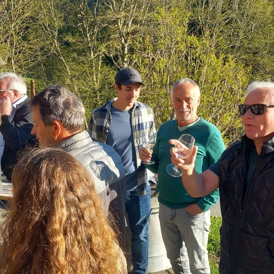 Mehrere Menschen stehen im Freien in einem bewaldeten Gebiet und unterhalten sich. Ein Mann hält ein Weinglas, andere sind in Freizeitkleidung. Im Hintergrund sind Bäume und Grün zu sehen.