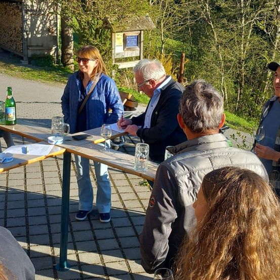Eine Gruppe von Menschen sitzt an einem Picknicktisch, möglicherweise auf einer Weinbergtour, wobei ein Mann auf Papier schreibt und eine Frau eine Weinflasche hält.