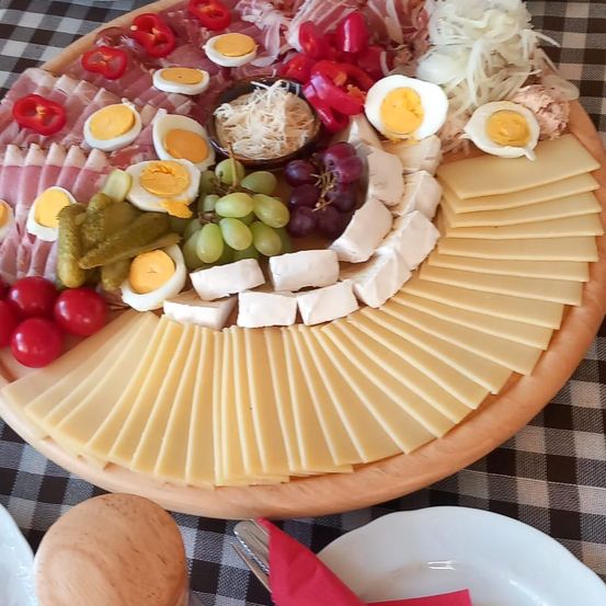 Ein Holztablett gefüllt mit einer Vielzahl von Lebensmitteln, darunter geschnittener Käse, geschnittene Eier, Trauben und Gurken. Es gibt auch Fleischscheiben und eine Schüssel mit geriebenem Käse.