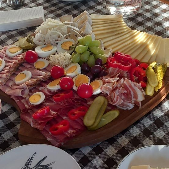 Ein hölzernes Tablett mit verschiedenen Fleischsorten, Käse, Eiern, Trauben, Tomaten und Gewürzgurken. Der Tisch ist mit einer karierten Tischdecke bedeckt.