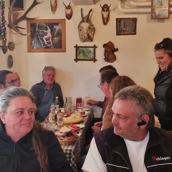 Eine Gruppe von Menschen sitzt in einem Restaurant. Ein Tisch mit Essen steht im Vordergrund. Einige Bilderrahmen sind an der Wand montiert. Eine Person steht neben dem Tisch.