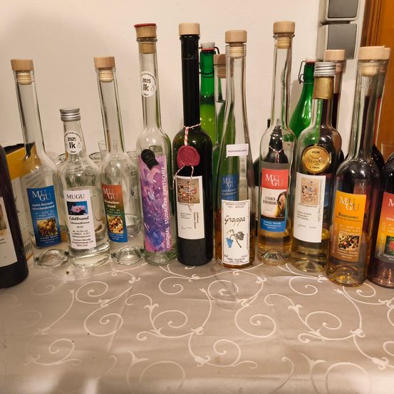 Ein Tisch hat mehrere Flaschen Alkohol mit Korken, darunter Wein, Grappa und Spirituosen, auf einem weißen Tuch. Die Flaschen haben weiße Etiketten mit Text und Bildern.