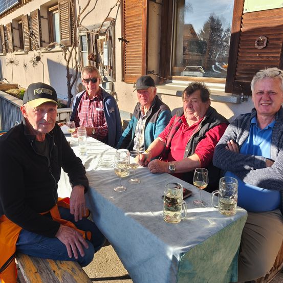 Fünf ältere Männer sitzen lächelnd um einen Tisch im Freien. Sie haben Weingläser und Bierkrüge. Der Tisch hat eine blaue Tischdecke. Sie sitzen vor einem Gebäude mit Fenstern und einem Baum.