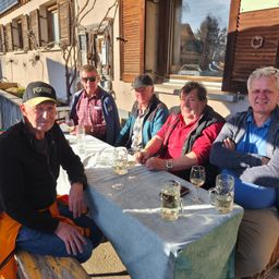 Fünf ältere Männer sitzen lächelnd um einen Tisch im Freien. Sie haben Weingläser und Bierkrüge. Der Tisch hat eine blaue Tischdecke. Sie sitzen vor einem Gebäude mit Fenstern und einem Baum.