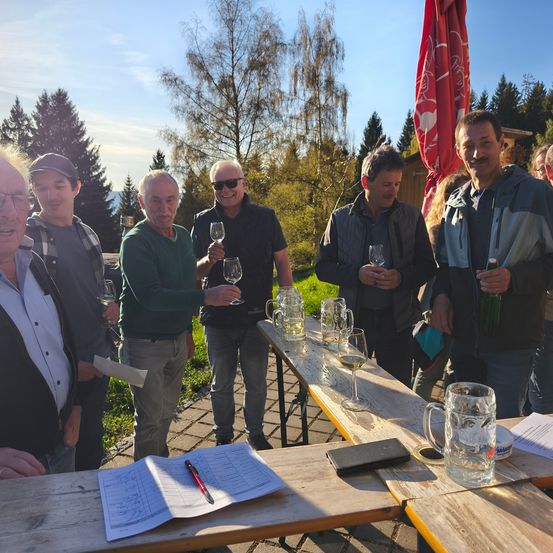 Eine Gruppe von Männern steht um Tische im Freien, hält Weingläser, lächelt und einer Mann hat ein Bier. Sie sind von Natur umgeben und es liegen Papiere, ein Notizbuch und ein Stift auf den Tischen.