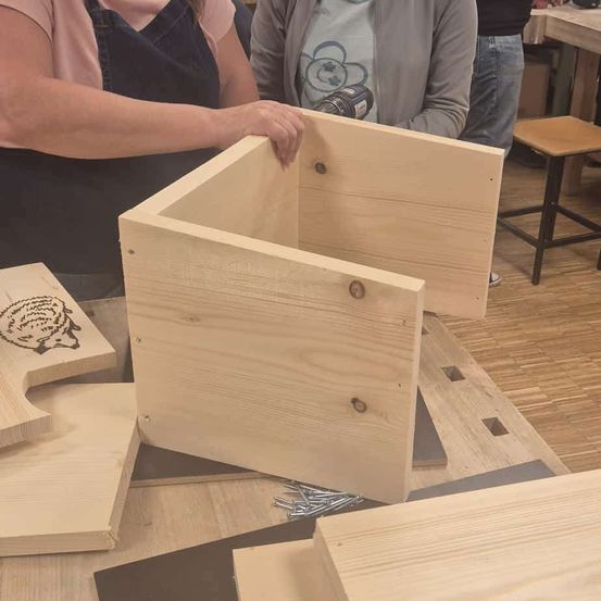 Zwei Personen arbeiten an einer Holzkiste mit Igeldesign, eine bohrt, die andere hält sie. Sie stehen in der Nähe eines Holztisches.