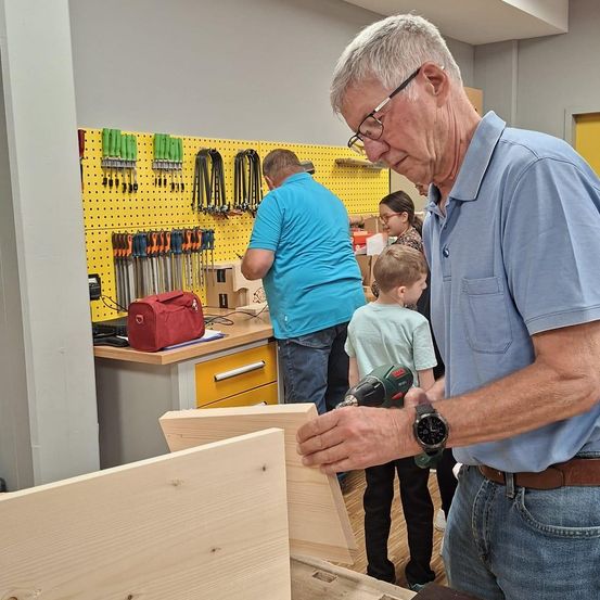 Ein älterer Mann benutzt einen Akkuschrauber, um in einer Werkstatt an einem Holzprojekt zu arbeiten. Andere Erwachsene und Kinder schauen zu. Werkzeuge hängen an einem gelben Lochblech.