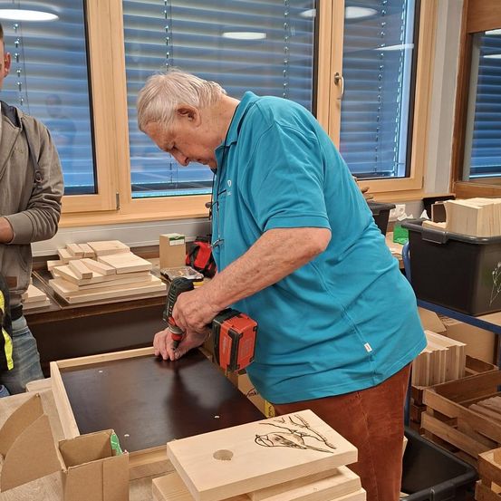 Ein älterer Mann in einem blauen Shirt und braunen Hosen benutzt einen Bohrer, um an einem Holzprojekt in einer Werkstatt mit Lagerboxen und Fenstern mit Jalousien zu arbeiten.