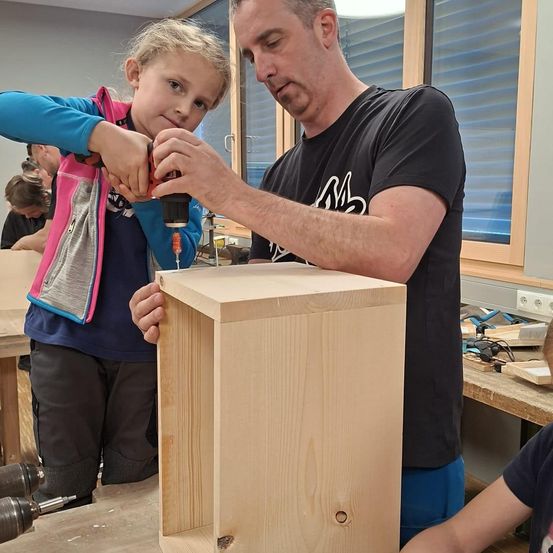 Ein Erwachsener und ein junges Mädchen sind in einer Werkstatt und arbeiten an einer Holzkiste. Das Mädchen hält eine Bohrmaschine, während der Mann zusieht. Hinter ihnen sitzt eine weitere Person. Die Werkstatt hat Holztische, Werkzeuge und ein Fenster.