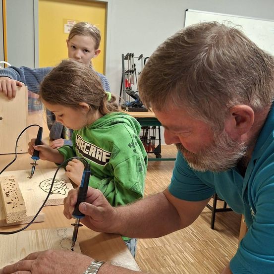 Ein Mann hilft zwei Kindern mit Holzbrandwerkzeugen, eines hält ein Werkzeug und das andere beobachtet aufmerksam. Sie sind in einer Werkstatt mit Holzplatten und Werkzeugen auf dem Tisch.