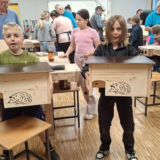Zwei Kinder halten Holzkästen mit Igel-Zeichnungen. Sie stehen in einer Werkstatt mit anderen, die arbeiten. Dahinter geht eine Frau in pinken Hosen. Ein Junge im grünen Shirt sitzt auf einem Stuhl.