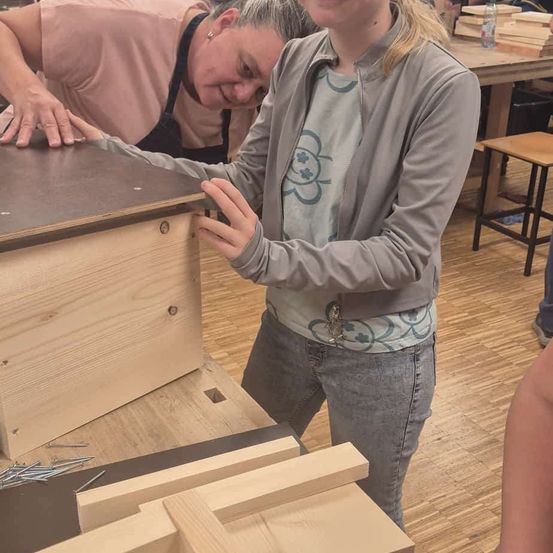 Eine Frau hilft einer anderen Frau bei der Arbeit an einer Holzkiste in einer Werkstatt. Die Frau trägt eine graue Jacke und Jeans. Die Holzkiste steht auf einem Holztisch mit einigen Nägeln.