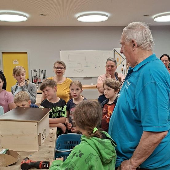 Eine Gruppe von Kindern und Erwachsenen versammelt sich in einer Werkstatt. Ein älterer Mann in einem blauen Hemd steht vor ihnen und spricht. Die Kinder schauen sich eine Holzkiste auf dem Tisch an.