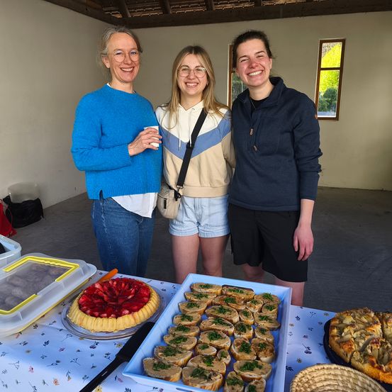 Drei Frauen stehen hinter einem Tisch mit Essen. Eine hält einen Becher. Sie lächeln und scheinen für ein Foto zu posieren. Es gibt mehrere Gerichte auf dem Tisch.