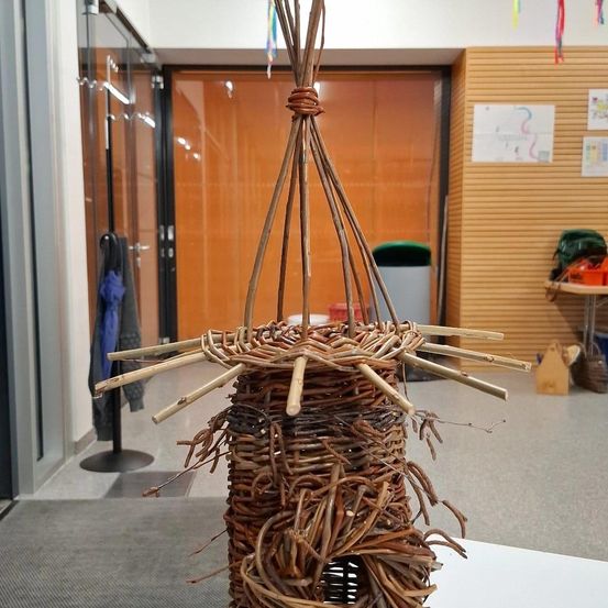 Ein handgefertigtes Vogelhaus aus Zweigen steht in einem Innenbereich. Es steht auf einer weißen Oberfläche in der Nähe einer Holzwand mit Glastüren und einem Mülleimer.