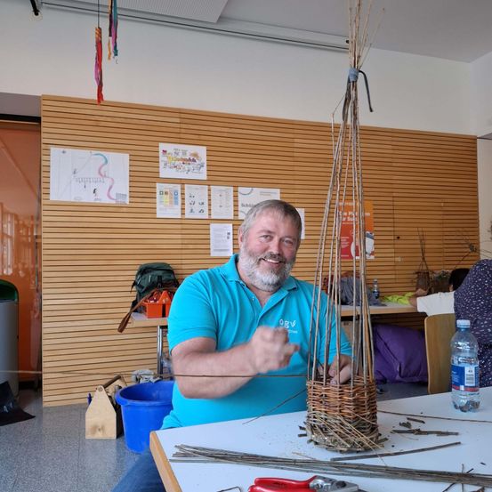 Ein Mann in einem blauen Shirt sitzt und arbeitet in einem Raum an einem Holzhandwerk.