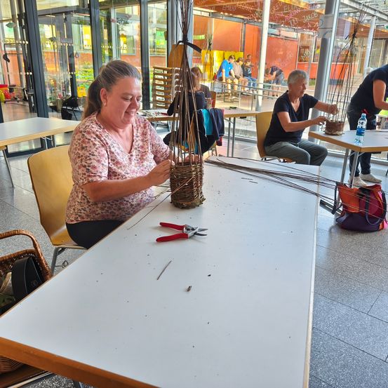 Eine Frau lächelt, während sie einen Korb aus Zweigen bastelt. Eine weitere Handwerkerin sitzt in der Nähe. Sie befinden sich in einem Raum mit Tischen, Stühlen und einer Glaswand, die eine Sitzgelegenheit im Freien zeigt.
