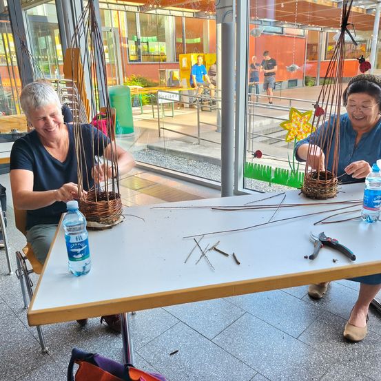 Zwei Frauen fertigen Körbe in einem Gebäude mit Glaswänden an. Ein Korb ist fertig, der andere wird bearbeitet. Ein Tisch hat eine Wasserflasche, Scheren und andere Werkzeuge. Menschen sind draußen zu sehen.