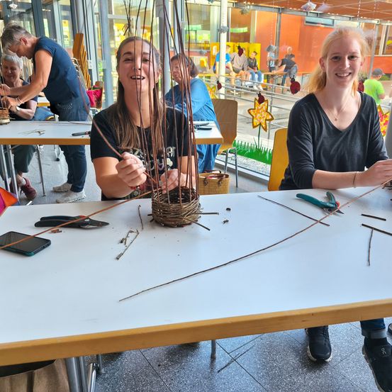 Zwei Mädchen basteln mit Zweigen in einer Werkstatt. Sie sitzen an einem Tisch, lächeln und haben einen Korb und Werkzeuge. Im Hintergrund sitzen und stehen Menschen an einer Glaswand.