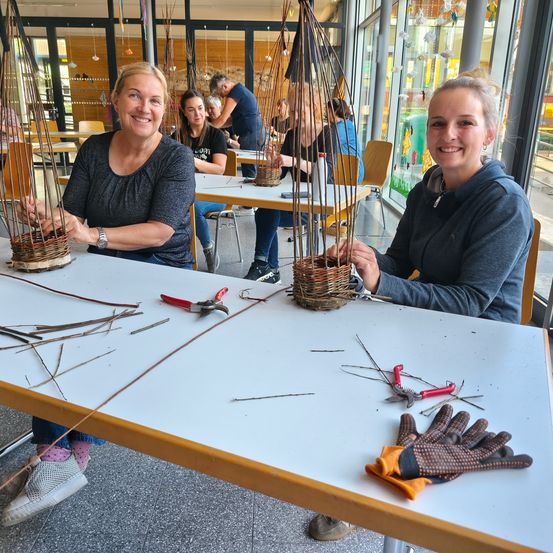 Zwei Frauen sitzen in einem Workshop an einem Tisch und halten Körbe aus Zweigen. Der Tisch ist mit Werkzeug, Handschuhen und weiteren Zweigen ausgestattet. Hinter ihnen arbeiten andere Personen an ähnlichen Projekten. Der Raum hat große Fenster und eine helle, luftige Atmosphäre.