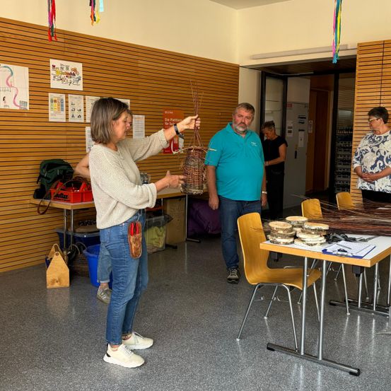 Eine Frau, die einen Korb hält, steht in einem Raum mit einem Mann, einem Tisch und einigen Stühlen. Im Hintergrund befinden sich eine Tür, Lagerboxen und Dekorationen.